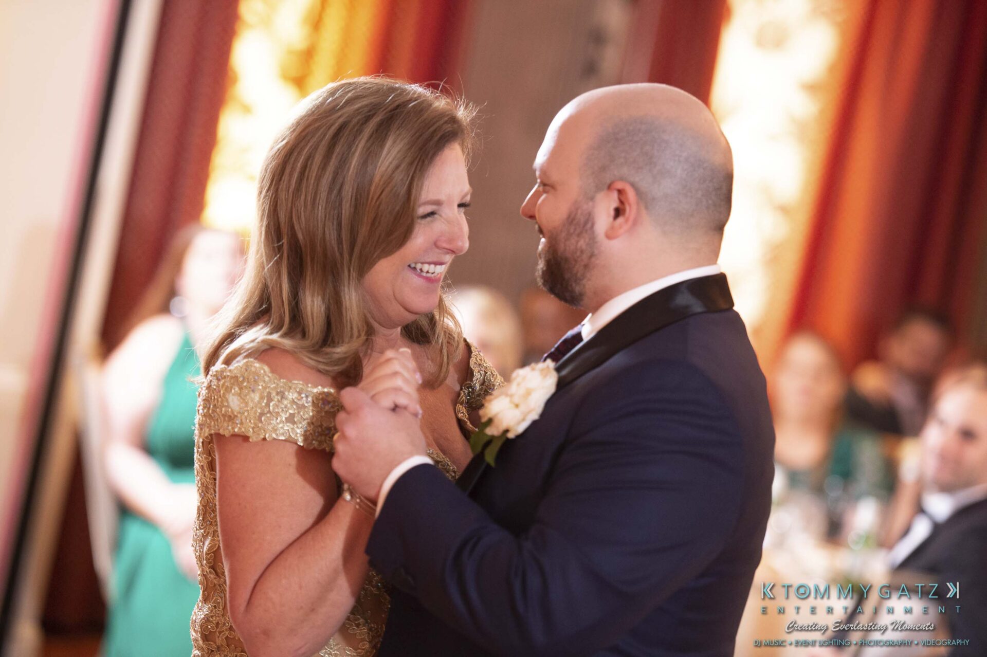 A couple dances together at a formal event, smiling and holding hands. The man wears a suit, and the woman is in a gold dress.