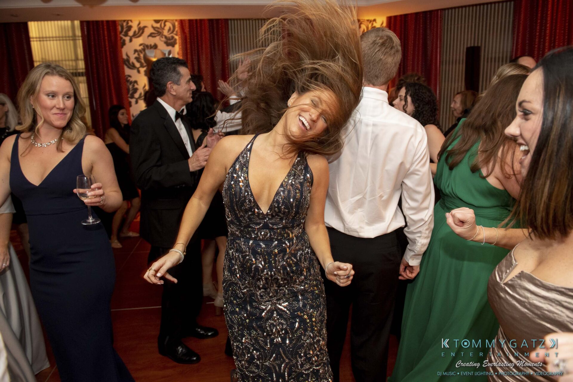 A woman in a sparkly dress dances energetically at a formal event, surrounded by other guests in evening attire.