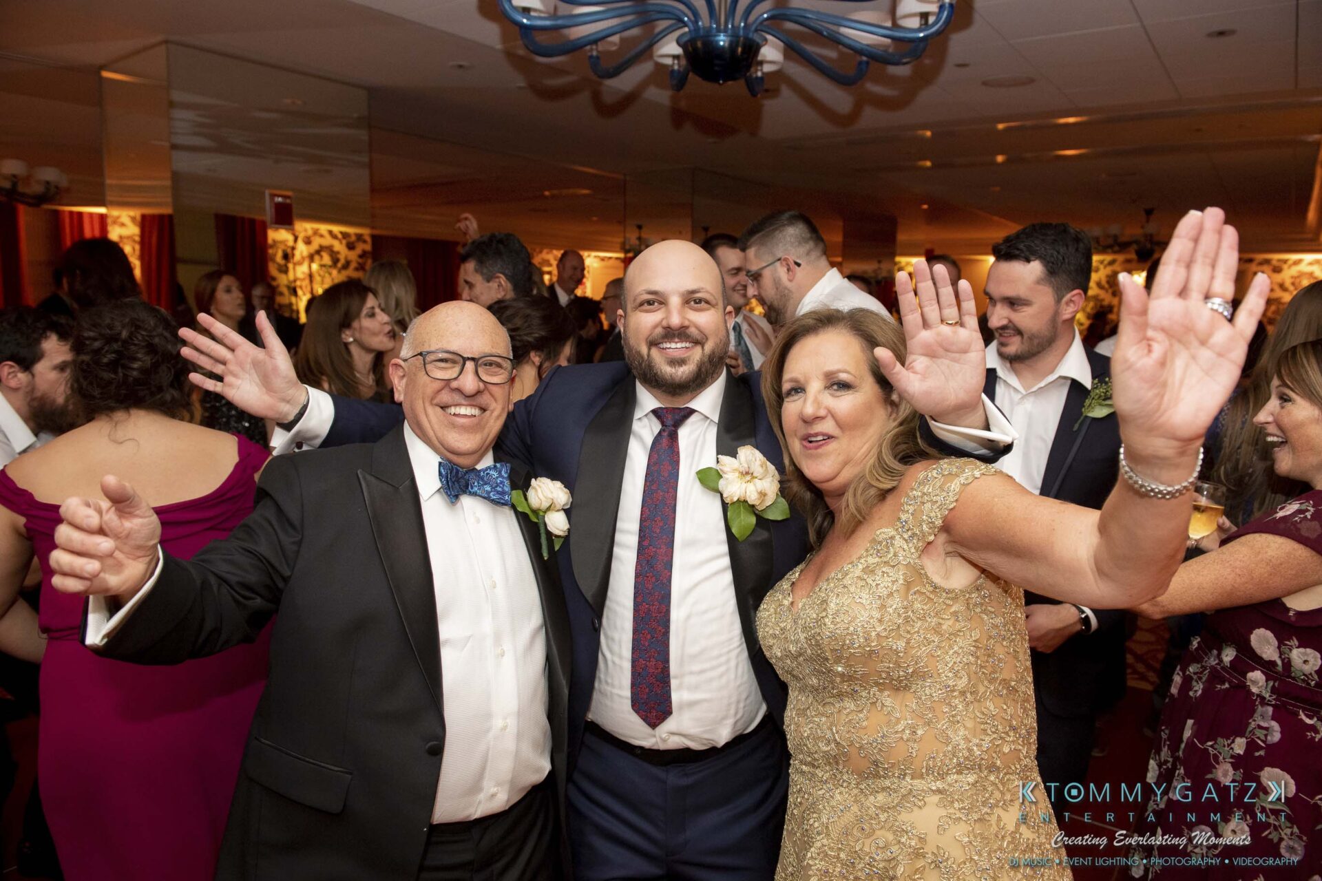 Three people smiling and posing with their arms up at a formal event, surrounded by other attendees.