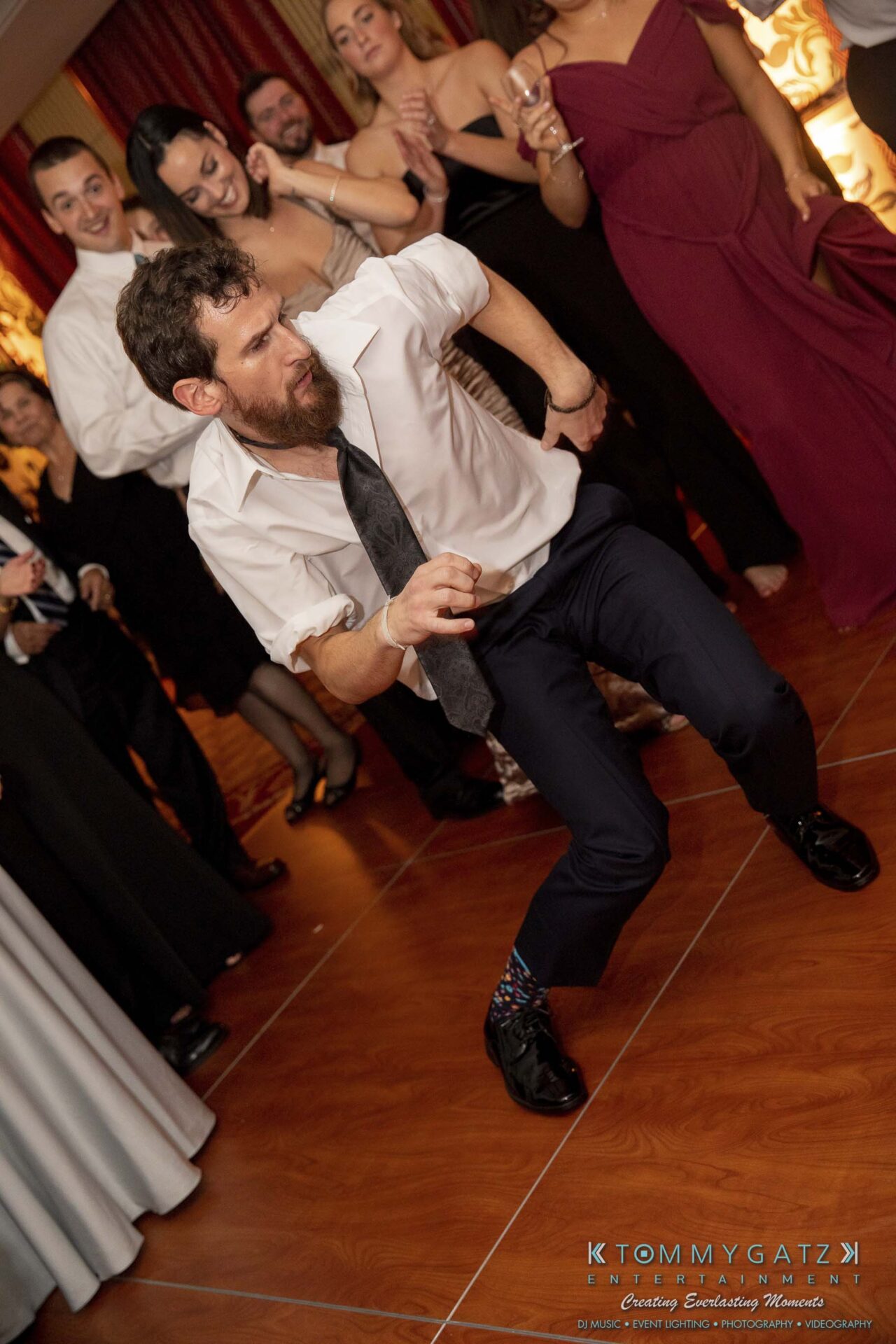 A bearded man in a white shirt and tie energetically dances on a wooden floor, surrounded by a group of people in formal attire at an indoor event.