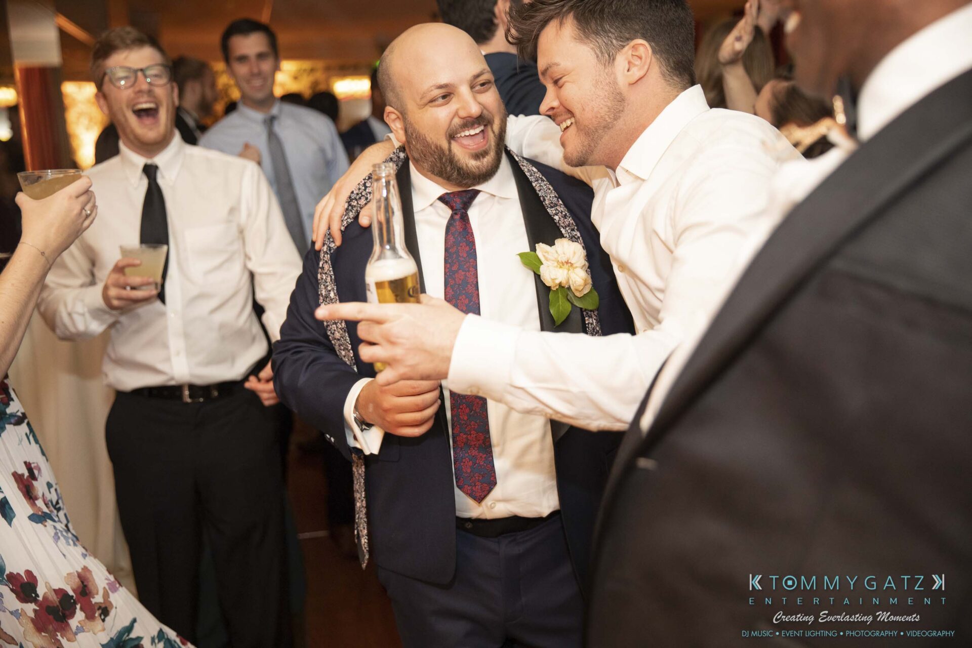 A man in a suit with a beer bottle smiles and embraces another man in a white shirt at a lively gathering. People in the background are dressed in formal attire, enjoying the event.