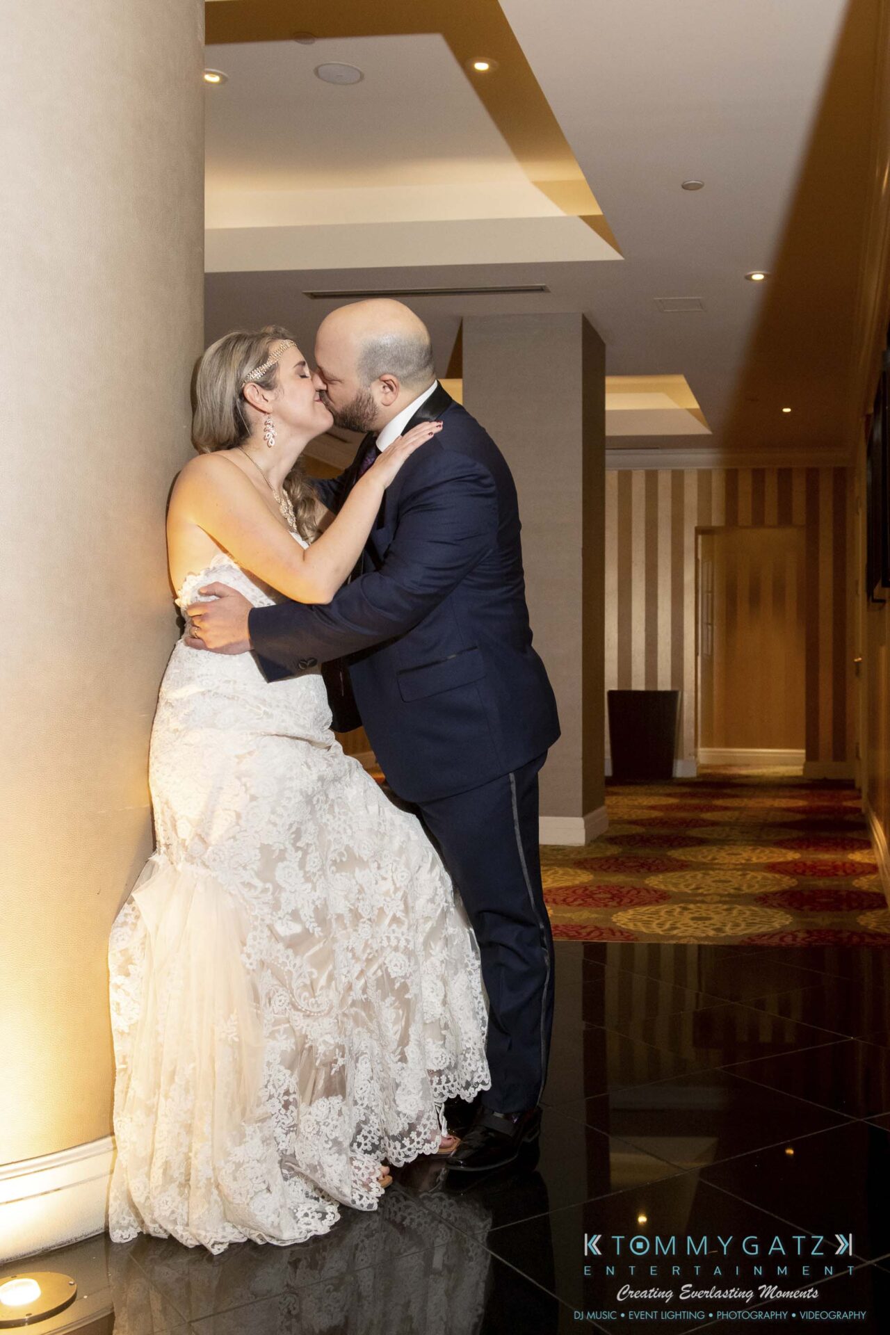 A bride and groom share a kiss in a dimly lit hallway adorned with striped wallpaper and patterned carpet.
