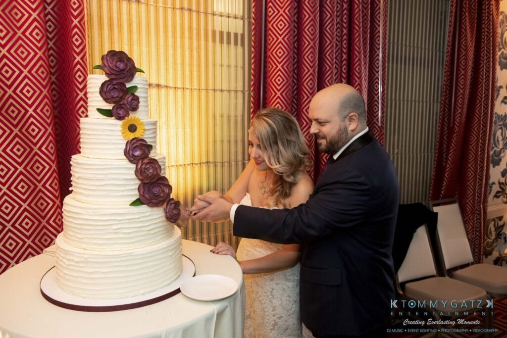A bride and groom cut a tiered white wedding cake adorned with purple flowers and sunflowers.