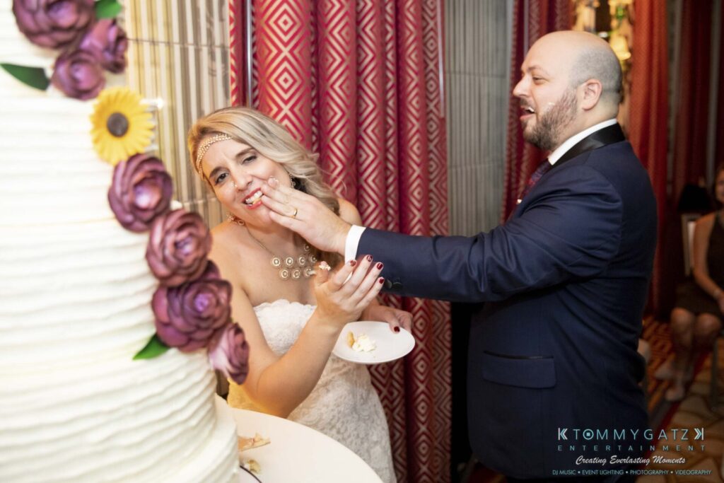 A bride and groom are playfully feeding each other cake in front of a decorated wedding cake with purple flowers.