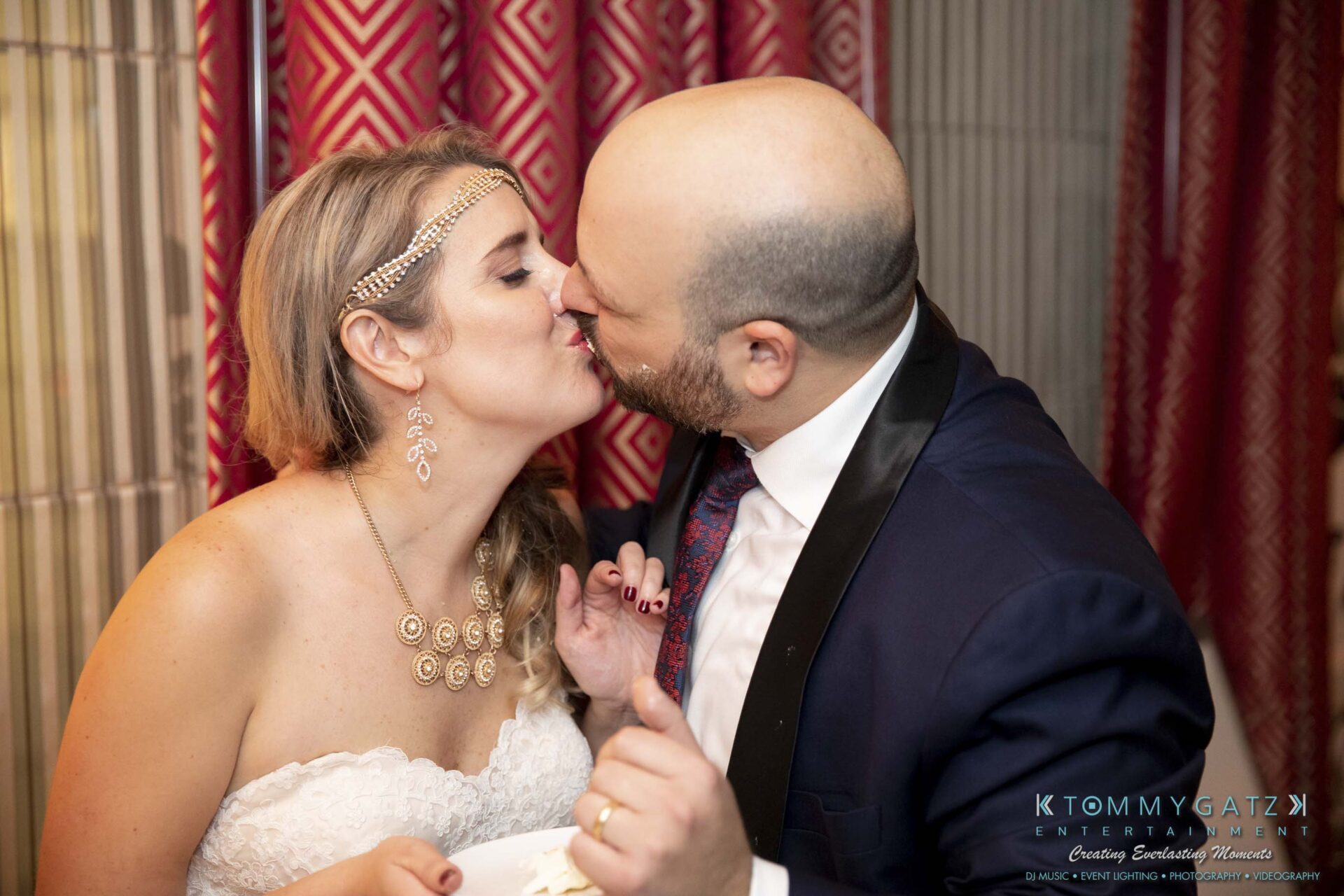 A bride and groom share a kiss during their wedding celebration, with the bride holding a piece of cake.