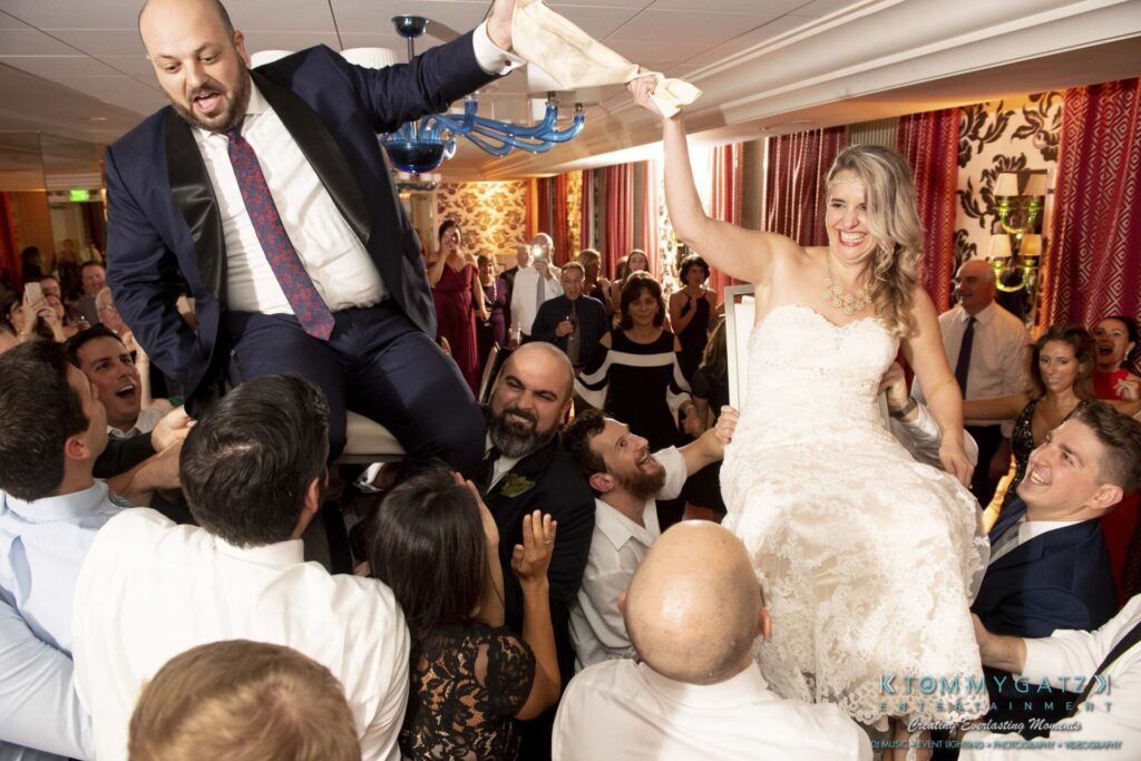 A wedding celebration with guests lifting a man and woman on chairs. The couple is smiling, and the crowd looks festive.