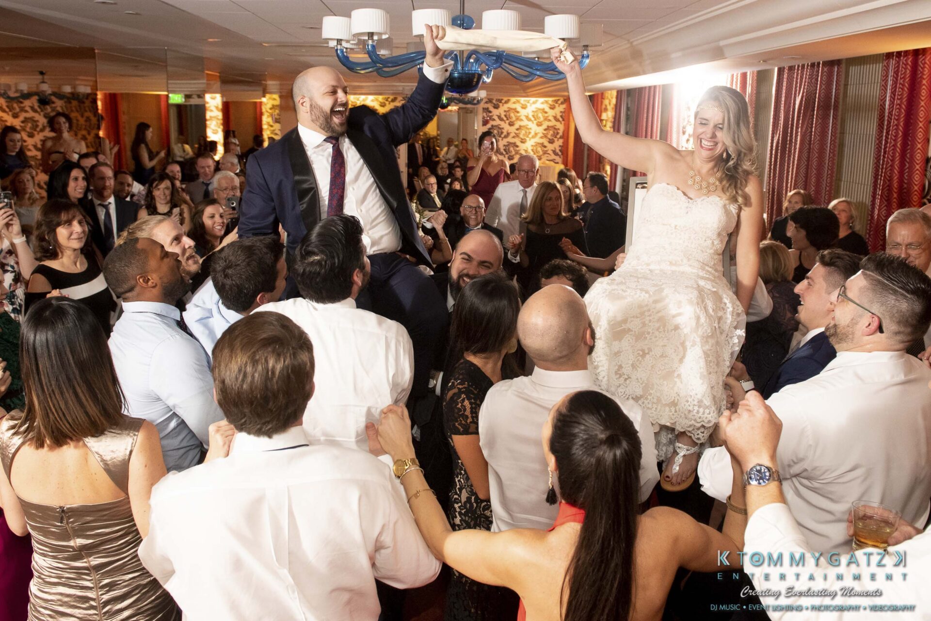 Bride and groom are lifted on chairs by guests at a lively indoor celebration. Guests are cheering and clapping around them.
