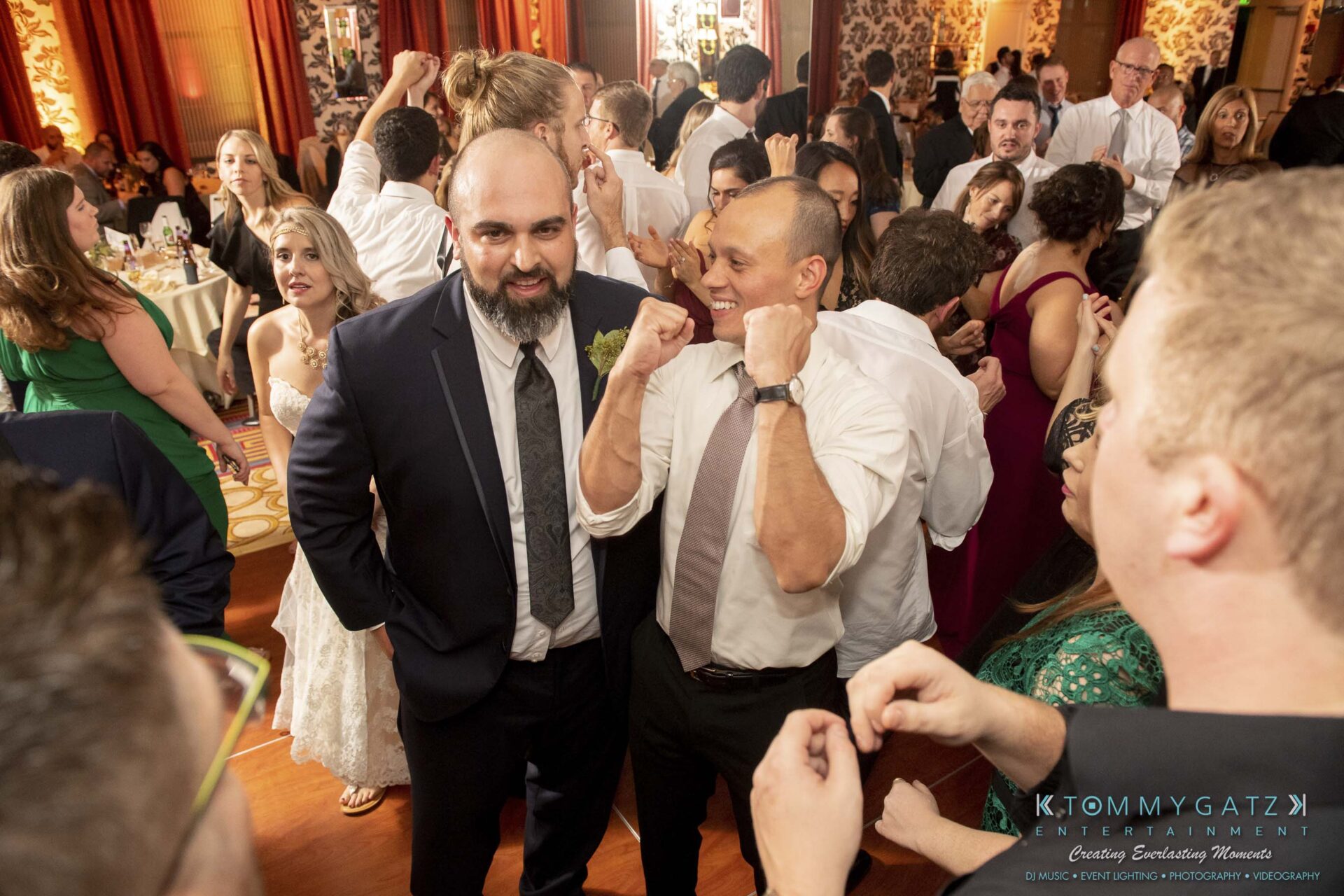 People dancing and celebrating at a formal event, with two men in suits smiling and posing with fists up in the foreground.