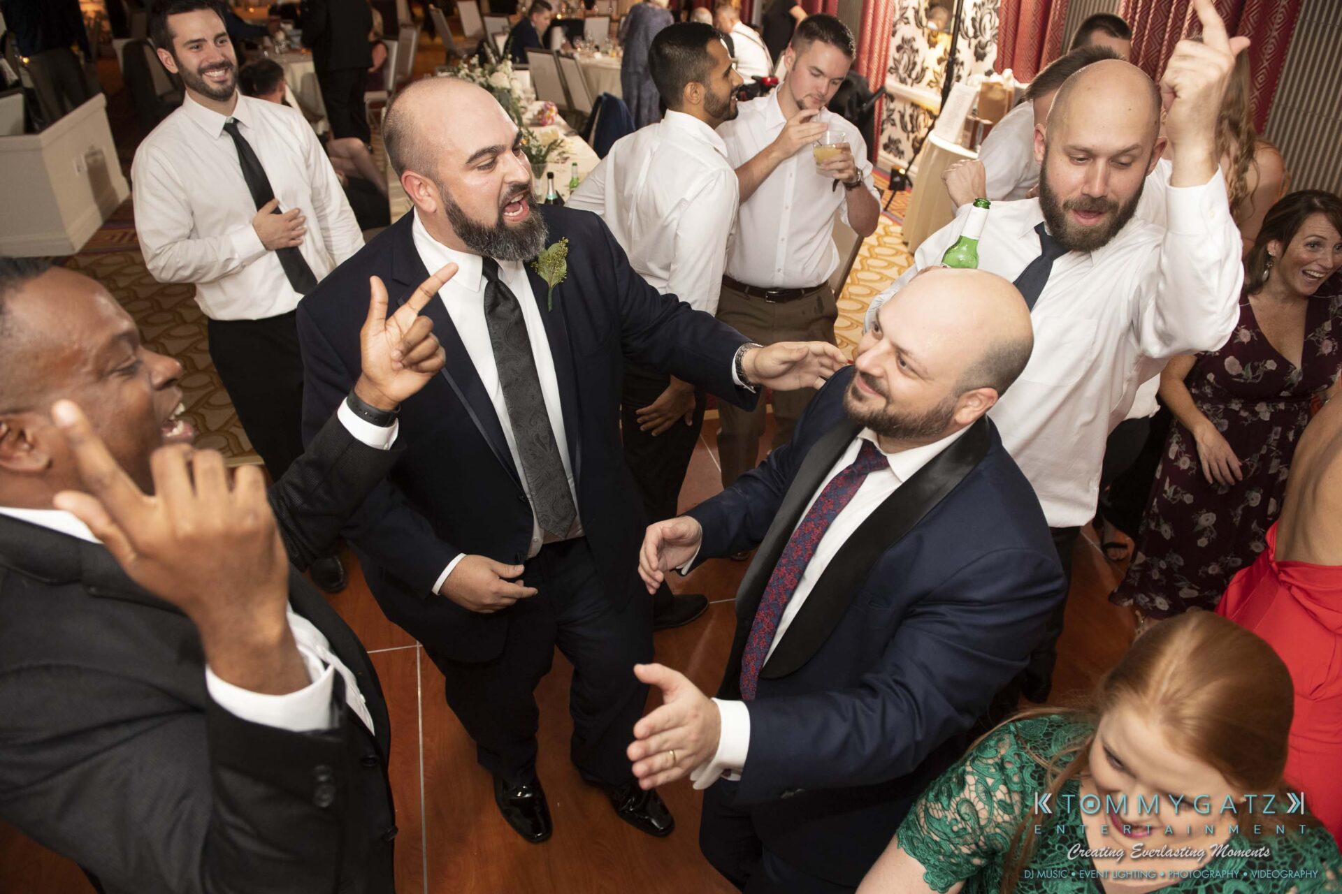 A group of people, mostly men in suits and ties, are joyfully dancing and raising their hands at an indoor event.