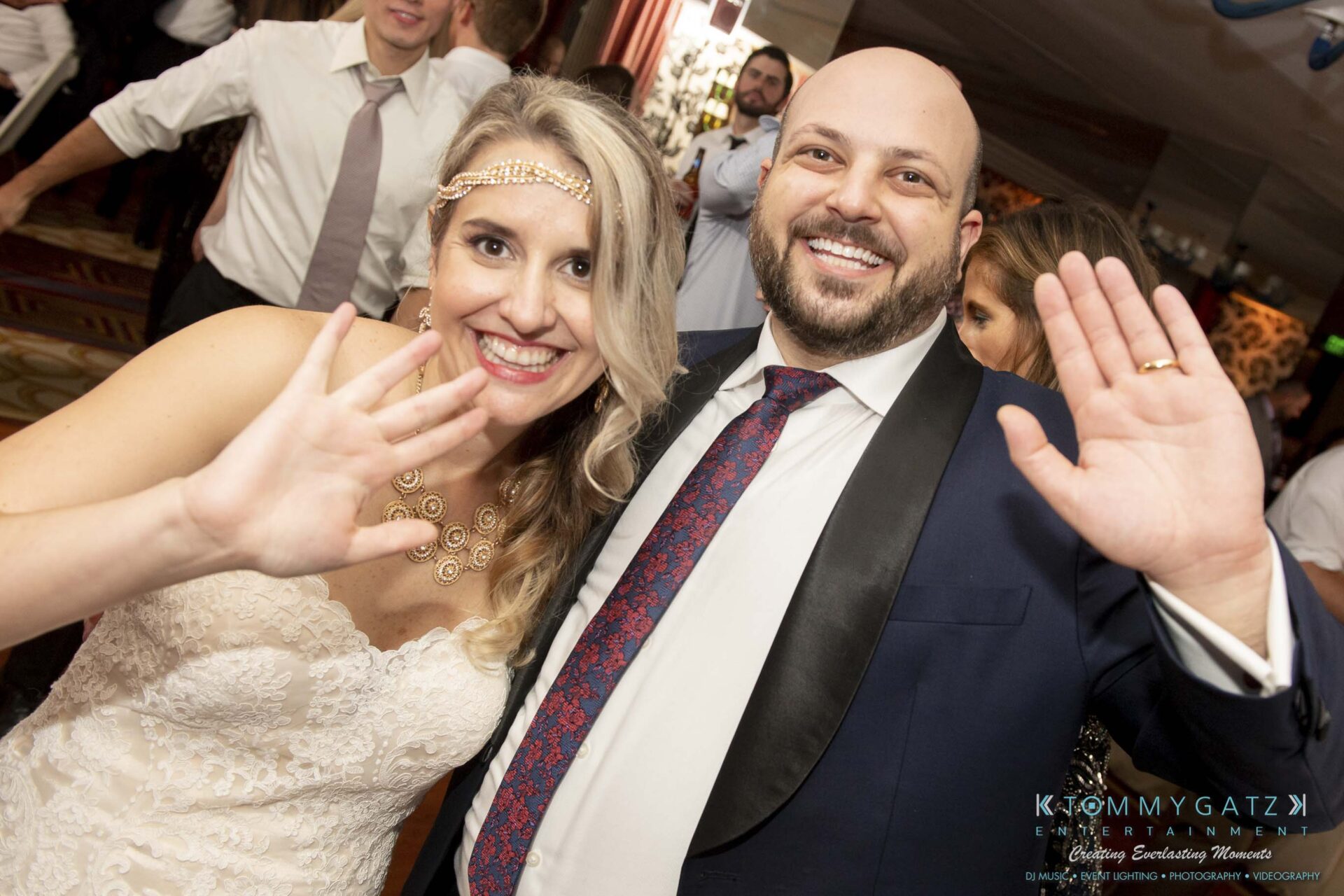 A smiling bride and groom wave at the camera during a celebration, surrounded by other guests in formal attire.
