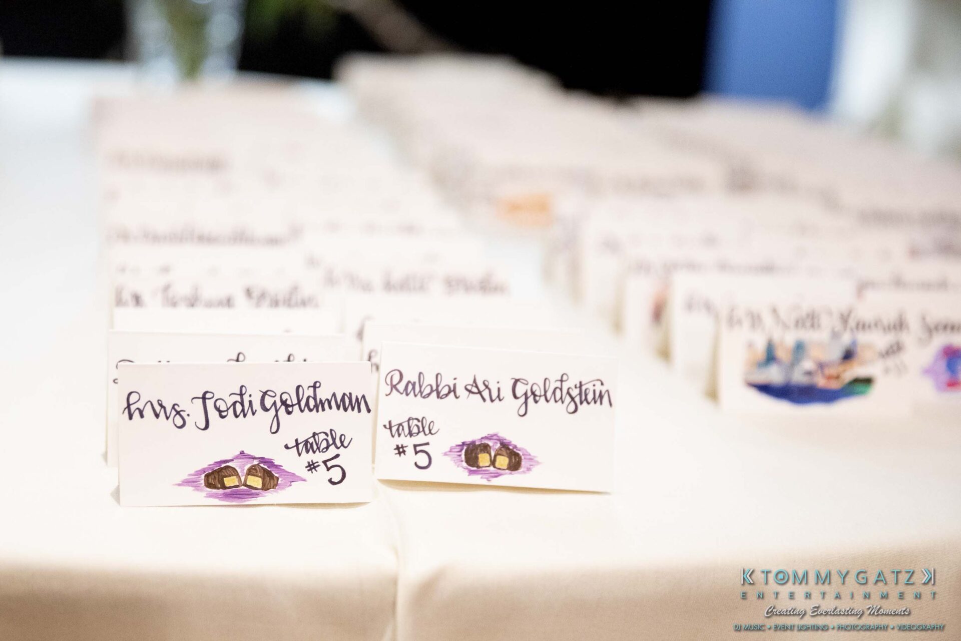 Place cards on a table, displaying names and table numbers, with one prominently reading "Rabbi Sri Goldstein.