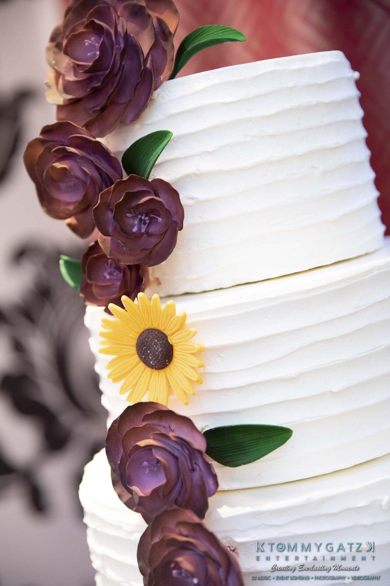 A three-tiered white cake decorated with purple roses and a yellow sunflower.