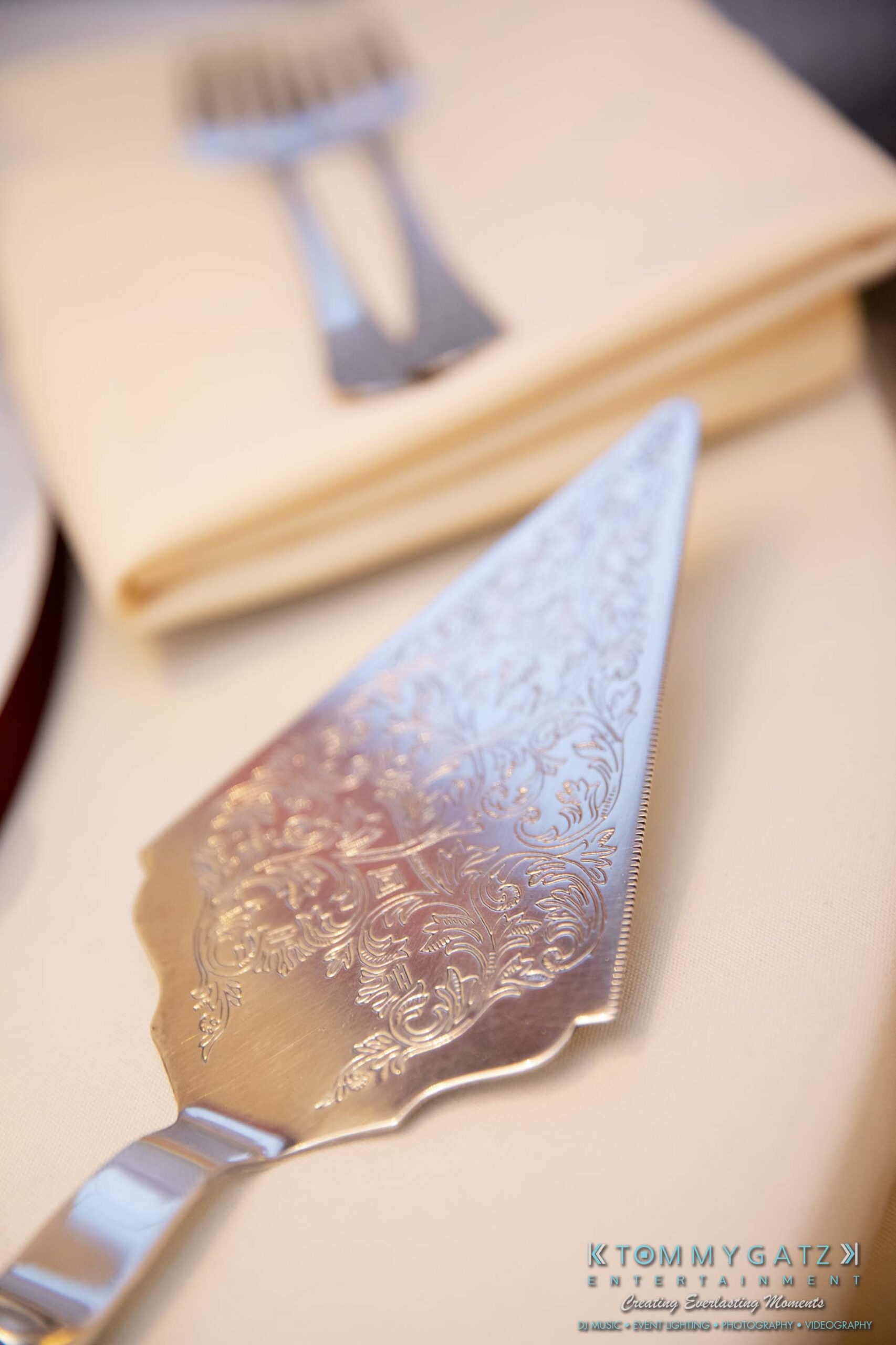 Close-up of an ornate silver cake server on a folded beige napkin, with the tines of a fork partially visible in the background.