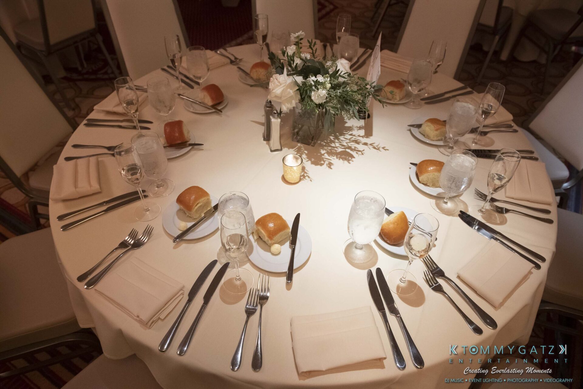 A round table set for a formal event with plates, rolls, glasses, cutlery, and a floral centerpiece.