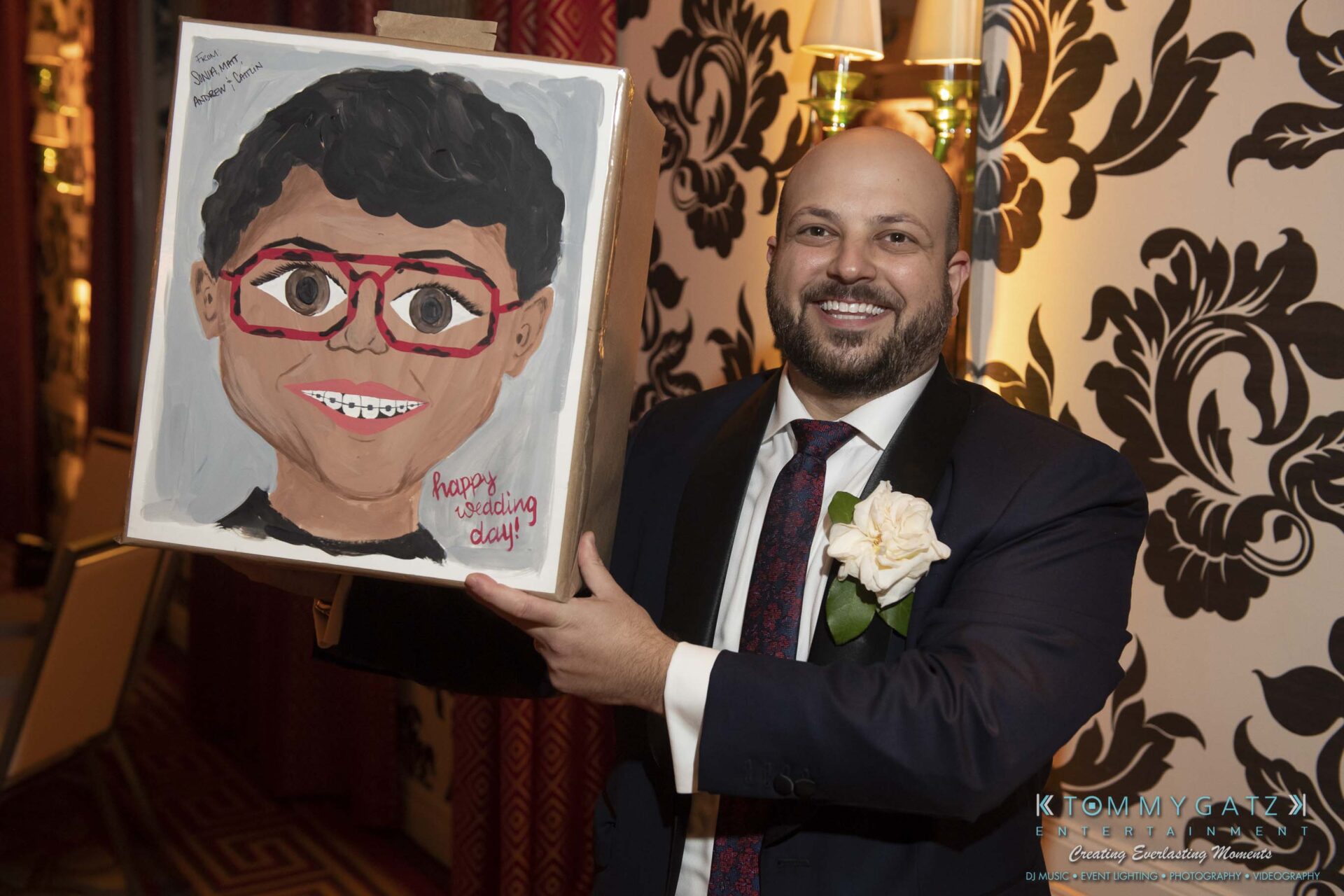 A smiling man in a suit holds a painting of a person with red glasses and "Happy Wedding Day" written on it.