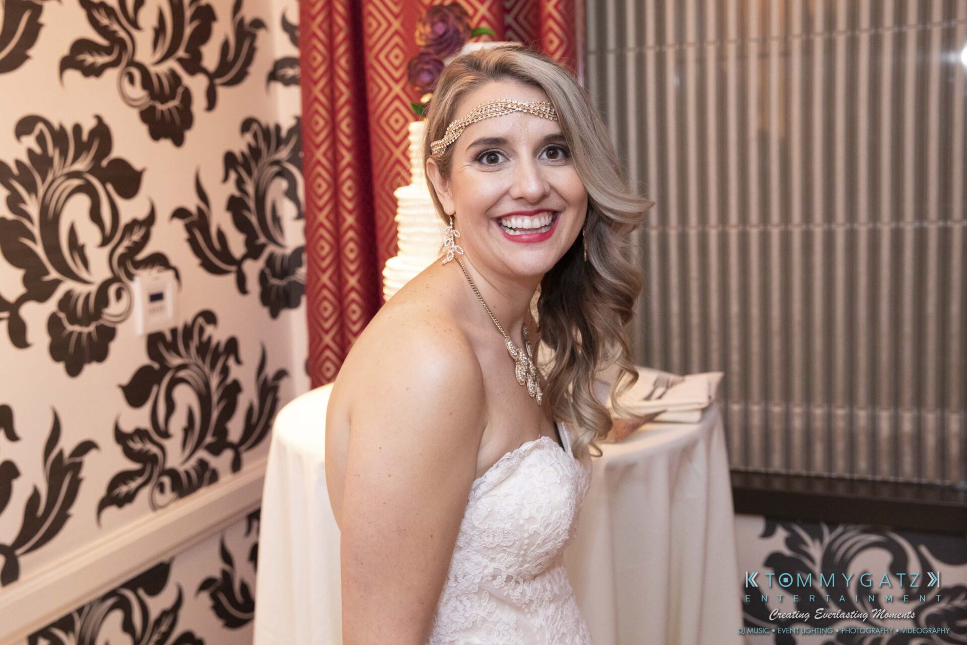 A woman in a strapless white dress smiles at the camera, standing in a room with patterned wallpaper and a small table in the background.