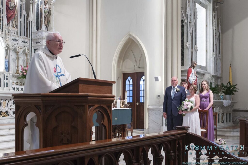 A priest speaks at a church altar. A couple stands nearby with two bridesmaids holding flowers. Marble columns and religious statues are in the background.