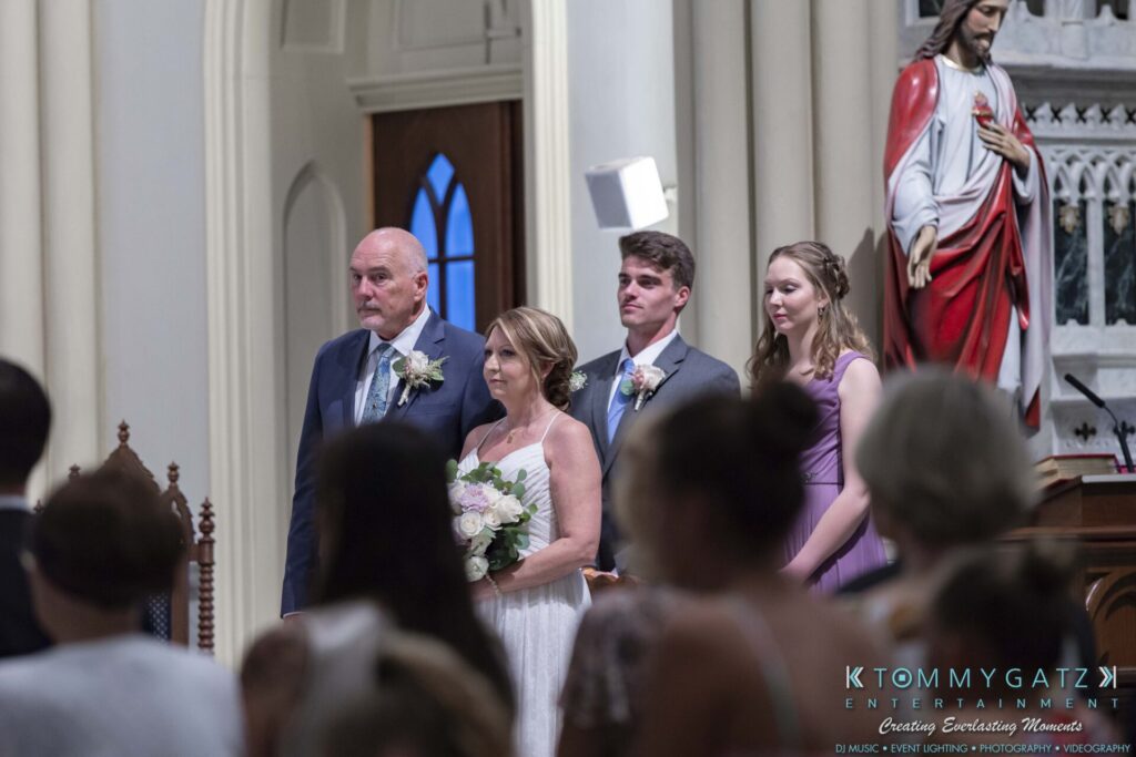 A wedding scene in a church with four people standing at the altar. The bride holds a bouquet, and a statue is visible in the background.