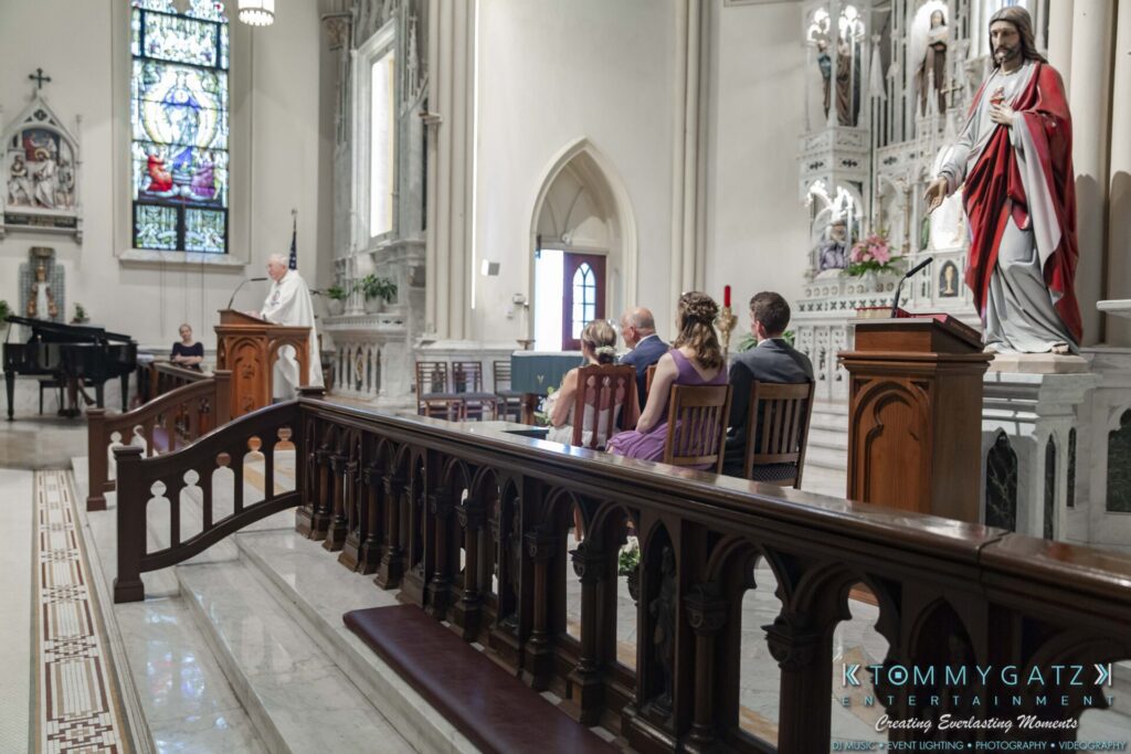 A priest speaks at an altar in a church while several people sit and listen. A statue of Jesus is visible nearby.