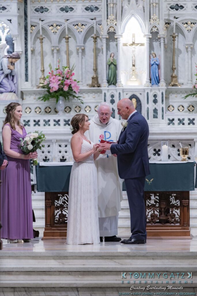 Grace-Michael-063018-CEREMONY-Low-Res-WM-133 - TOMMY GATZ ENTERTAINMENT A couple exchanges rings during a church wedding ceremony. A priest oversees the event, and two bridesmaids stand nearby.