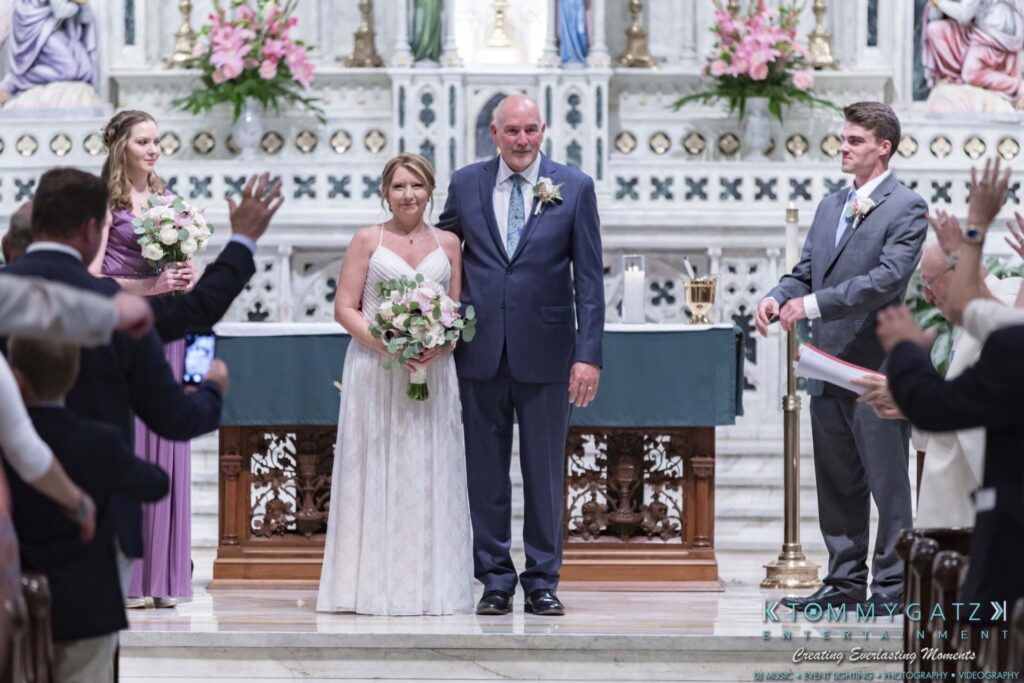 A bride and groom stand at the altar, holding bouquets, surrounded by wedding attendants and guests in a church setting.