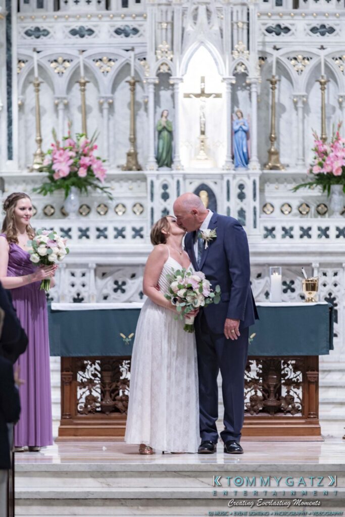 A bride and groom kiss at the altar during their wedding ceremony, with a bridesmaid holding flowers nearby.