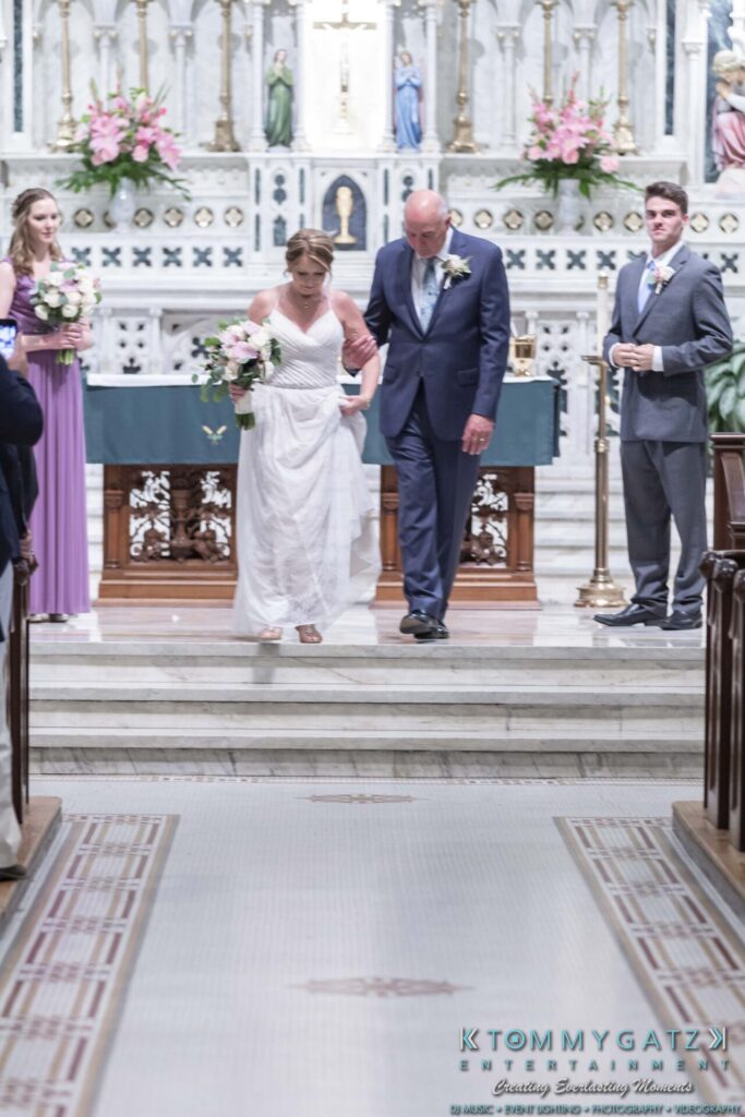 A bride and groom walk down the aisle in a church, accompanied by their wedding party, with floral arrangements in the background.