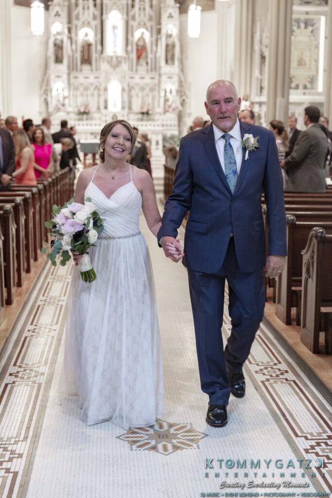 Grace-Michael-063018-CEREMONY-Low-Res-WM-174 - TOMMY GATZ ENTERTAINMENT A bride and groom hold hands, walking down the aisle in a church. The bride wears a white dress and holds a bouquet; the groom wears a blue suit with a boutonniere.