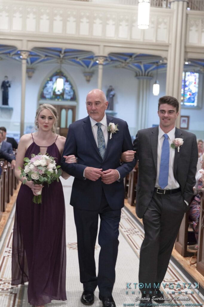 Grace-Michael-063018-CEREMONY-Low-Res-WM-59 - TOMMY GATZ ENTERTAINMENT Three people walking down an aisle in a church, with the two individuals on the sides wearing formal attire and holding flowers, and the person in the center wearing a suit.