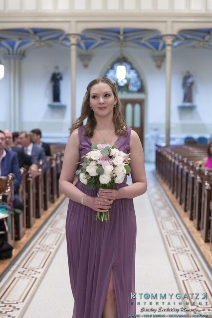 A woman in a purple dress holds a bouquet while walking down an aisle in a church.