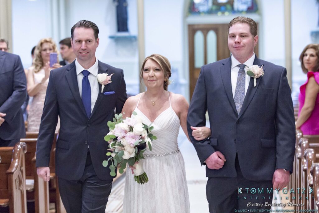 Grace-Michael-063018-CEREMONY-Low-Res-WM-74 - TOMMY GATZ ENTERTAINMENT A woman in a white dress walks down the aisle with two men in suits, one holding flowers. Guests are seated in the background.