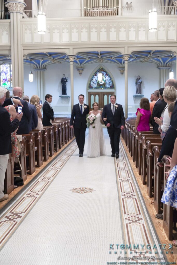 Bride walking down the aisle with two men in a church, surrounded by seated guests.