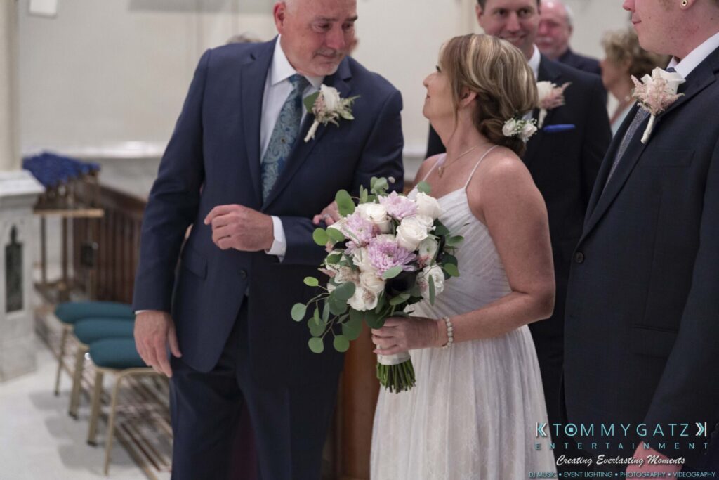 A bride holding a bouquet stands next to a man in a suit with a boutonniere. They are in a decorated indoor setting with chairs and flowers visible.