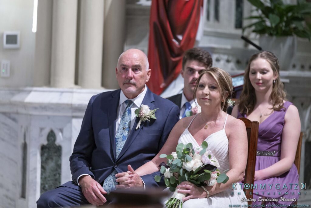 A bride and groom sit together, holding hands, during a wedding ceremony. They are accompanied by two attendants in formal attire.