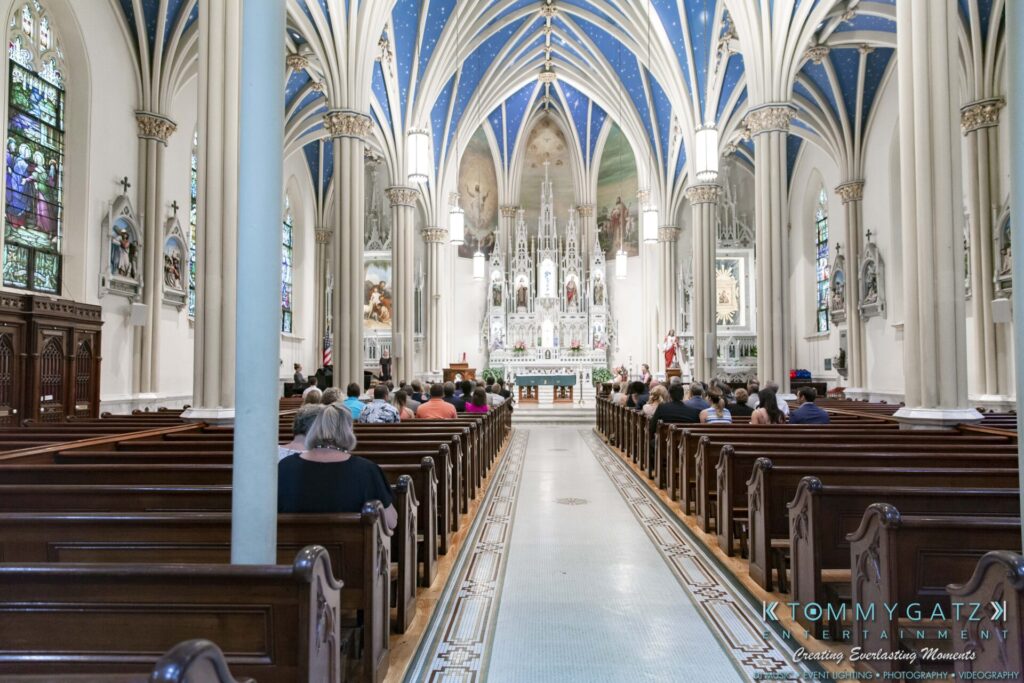 Grace-Michael-063018-CEREMONY-Low-Res-WM-96 - TOMMY GATZ ENTERTAINMENT Interior of a large church with people seated in wooden pews, facing an ornate altar. White columns and blue vaulted ceilings add to the cathedral's grand architecture.