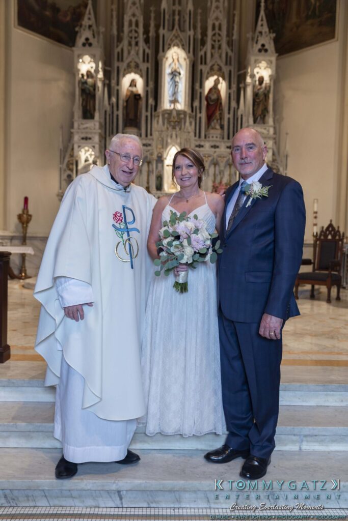 A bride and groom pose with a priest in a church. The bride holds a bouquet, and all are smiling.