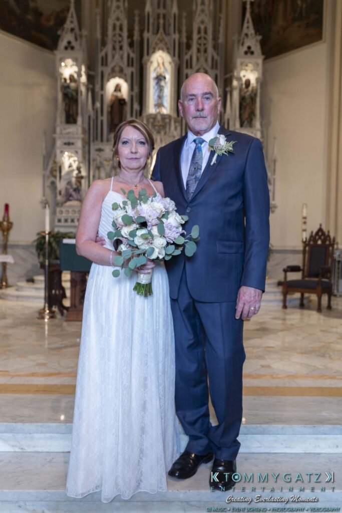 Grace-Michael-063018-FORMALS-Low-Res-WM-197 - TOMMY GATZ ENTERTAINMENT A couple in formal attire pose together in a church setting; the woman holds a bouquet, and the man wears a blue suit with a boutonniere.