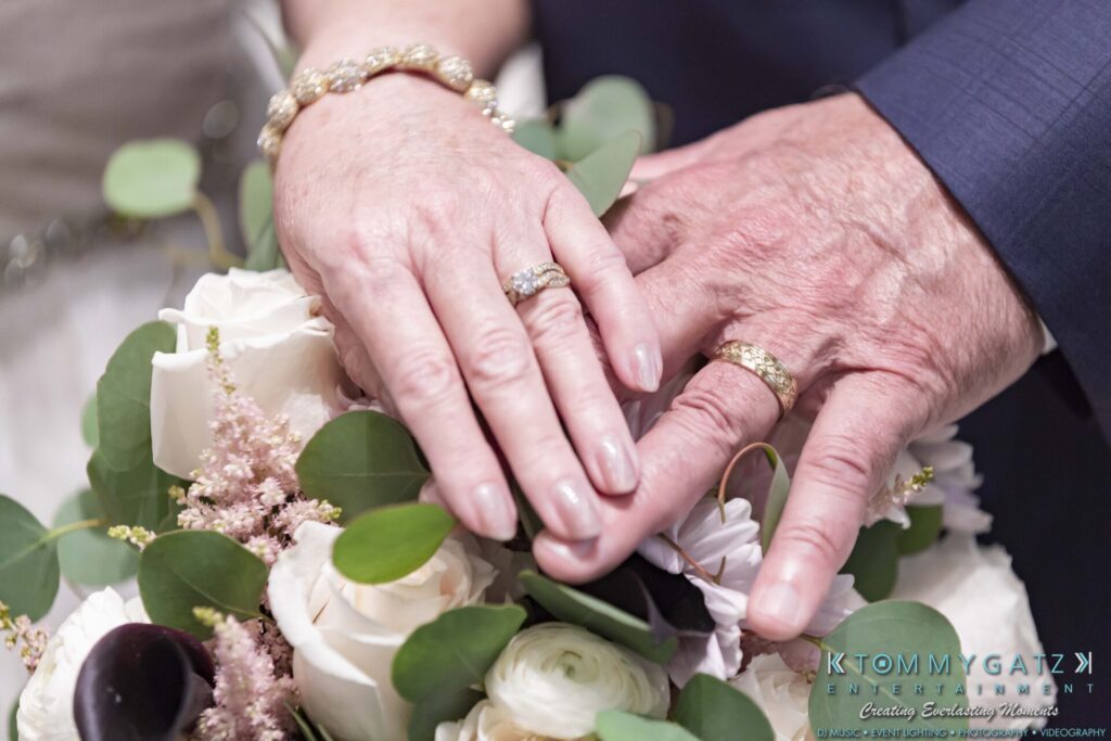 Close-up of two hands wearing wedding rings, resting on a bouquet of white and pink flowers with greenery.