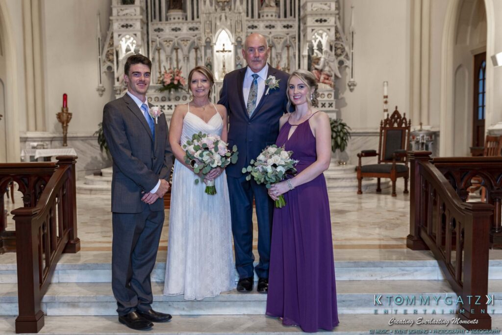 Grace-Michael-063018-FORMALS-Low-Res-WM-208 - TOMMY GATZ ENTERTAINMENT A wedding group photo in a church with stained glass and ornate architecture; two women holding bouquets, standing beside two men on a stone step.
