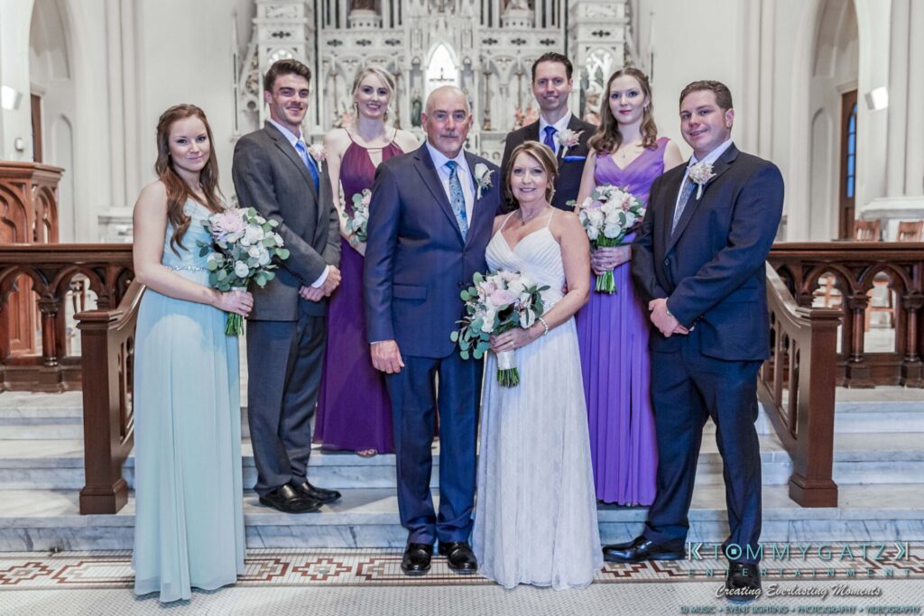 Grace-Michael-063018-FORMALS-Low-Res-WM-258 - TOMMY GATZ ENTERTAINMENT A wedding party poses in a church. The bride and groom stand in the center, surrounded by bridesmaids in colorful dresses and groomsmen in suits, all holding floral bouquets.