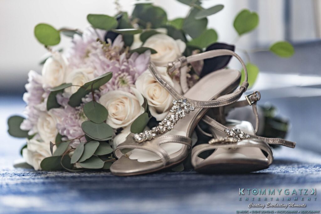 Grace-Michael-063018-PREP-Low-Res-WM-1 - TOMMY GATZ ENTERTAINMENT A bouquet of white and light purple flowers with green leaves sits next to a pair of jeweled, silver high-heeled sandals on a blue surface.