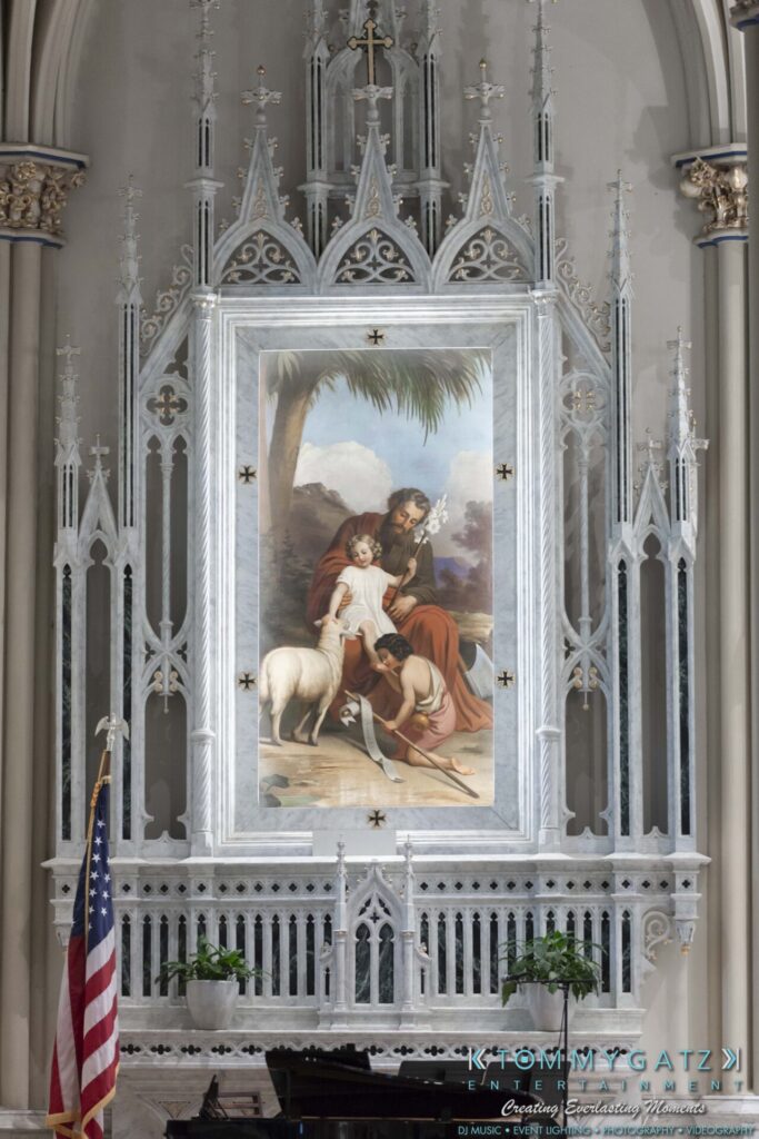 Grace-Michael-063018-PREP-Low-Res-WM-25 - TOMMY GATZ ENTERTAINMENT A large, ornate altar featuring a framed painting of religious figures with a lamb. An American flag stands on the left side.