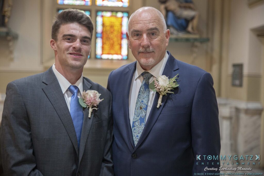 Grace-Michael-063018-PREP-Low-Res-WM-33 - TOMMY GATZ ENTERTAINMENT Two men in suits with boutonnières stand in a decorated indoor setting, featuring stained glass in the background.