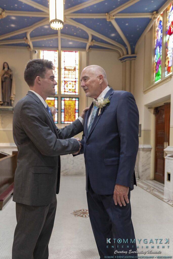 Two men in suits stand in a church, shaking hands. Stained glass windows and religious statues are visible in the background.