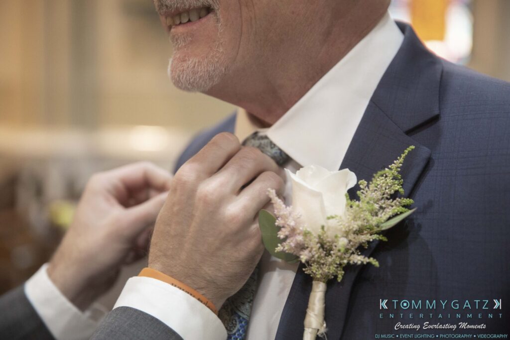 A person adjusts an older man's tie, who is wearing a suit with a boutonniere, during an indoor event.