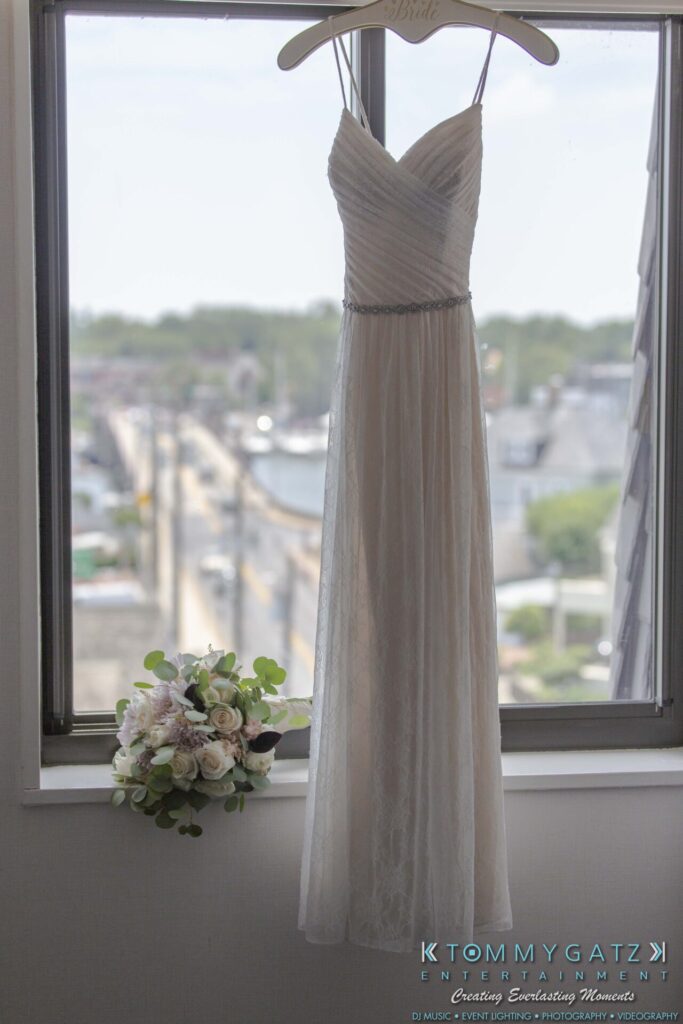 A white wedding dress hangs in front of a window, with a bouquet of pale flowers on the windowsill.