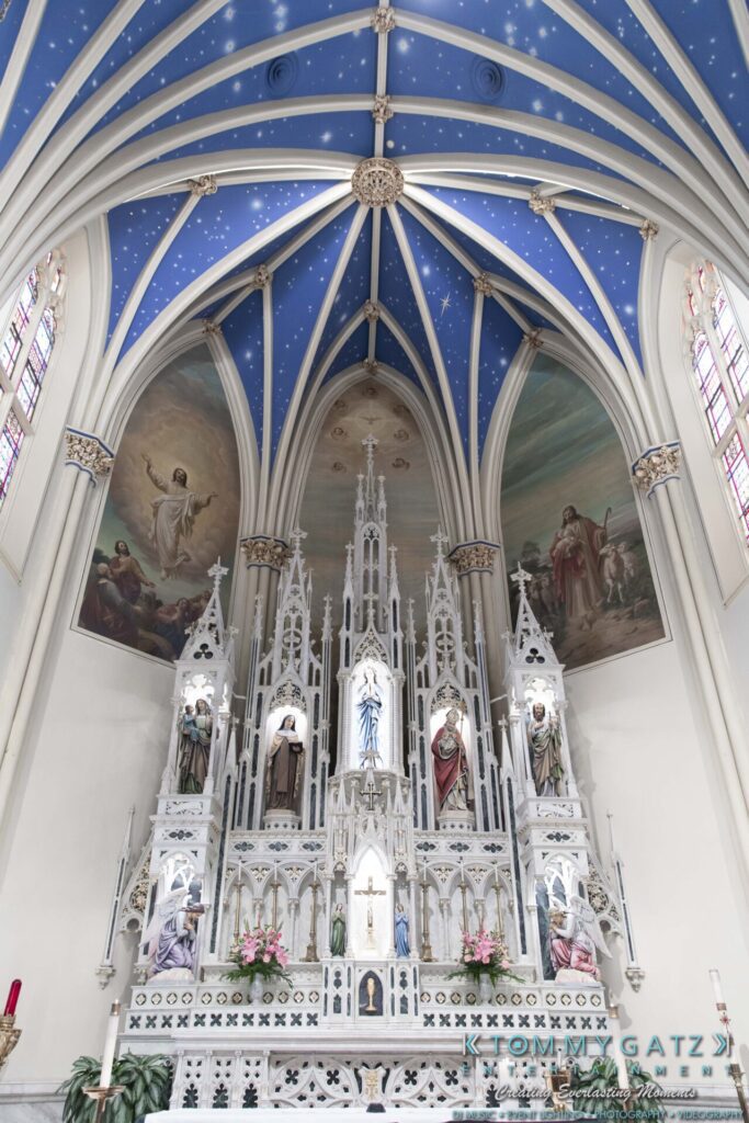 Grace-Michael-063018-PREP-Low-Res-WM-55 - TOMMY GATZ ENTERTAINMENT Gothic church interior with a blue, starry vaulted ceiling and ornate white altar featuring religious statues and stained glass windows.