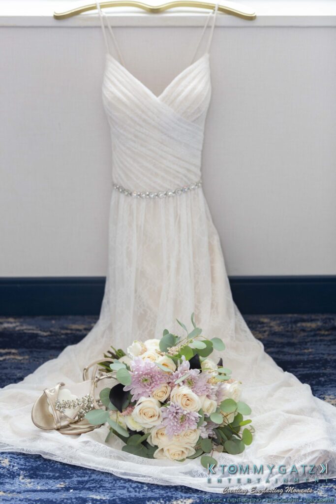 A white wedding dress with gem detailing is displayed against a wall. In front, there's a bouquet of pink and white flowers, and a pair of silver shoes on a blue carpet.