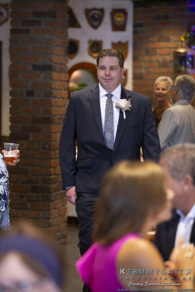 A man in a suit with a flower boutonniere walks through a room with people and ornamental wall decor in the background.
