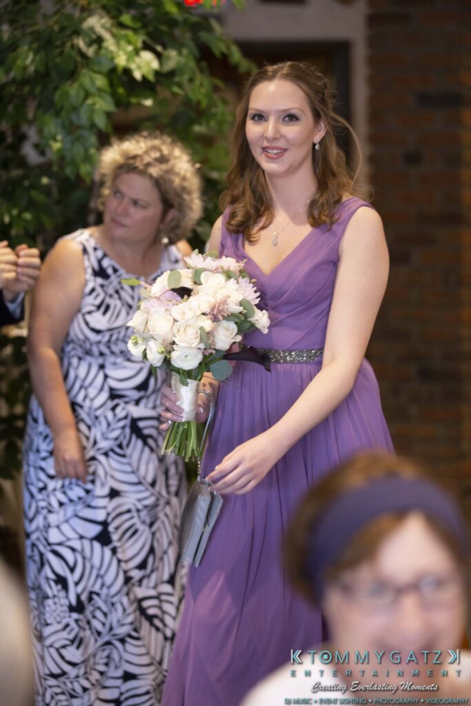 Grace-Michael-063018-RECEPTION-Low-Res-WM-274 - TOMMY GATZ ENTERTAINMENT A woman in a purple dress holds a bouquet while smiling. Another woman in a patterned dress stands behind her.